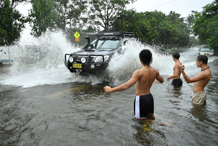 Three boys wading in floodwater direct a car through a suburban street