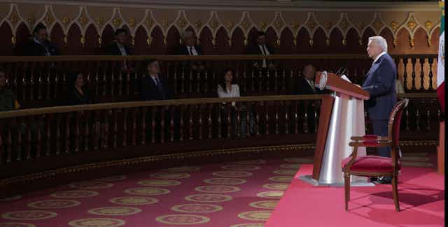 El presidente de México, Andrés Manuel López Obrador. lee un discurso ante una audiencia que escucha entre sombras.
