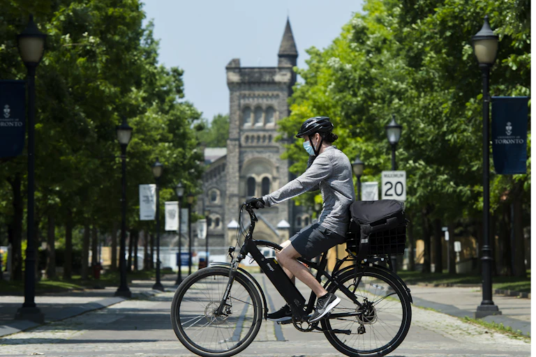 A person wearing a mask and a bike helmet cycles past the University of Toronto, with trees in the background.