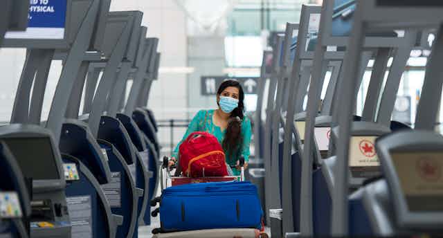 A woman with long dark hair wearing a mask pushes her luggage cart between rows of empty check-in kiosks at Toronto's Pearson airport.