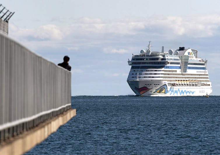 A huge cruise ship approaches a harbour as a person watches at the end of a pier in the foreground.