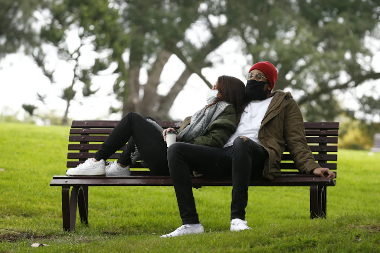 A man and a woman, wearing masks, sit on a bench