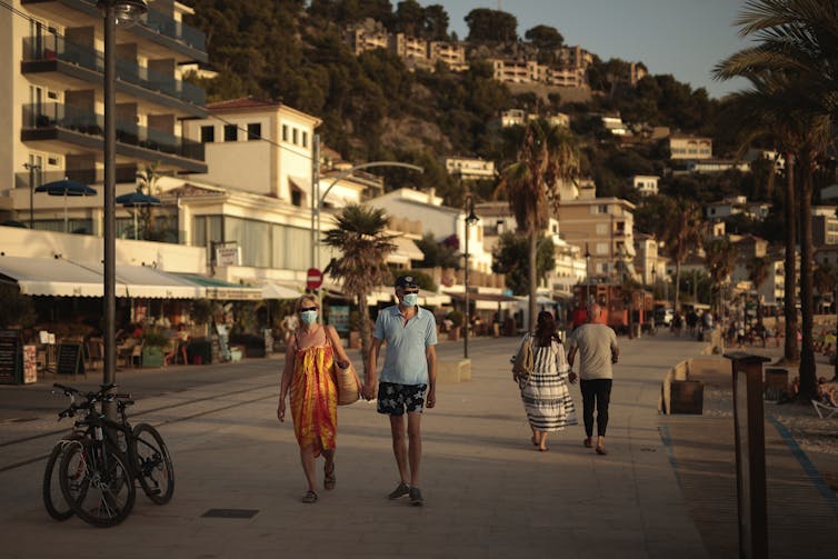 A couple holds hands as they walk along a promenade, with white-washed buildings behind them and palm trees to the right.