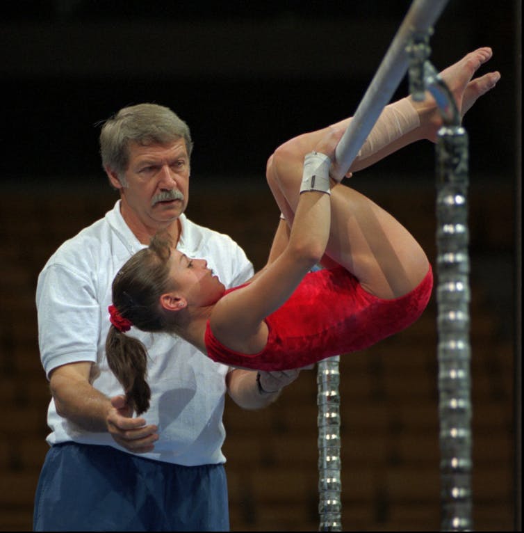 Young gymnast hangs on a bar, watched over by an older man.