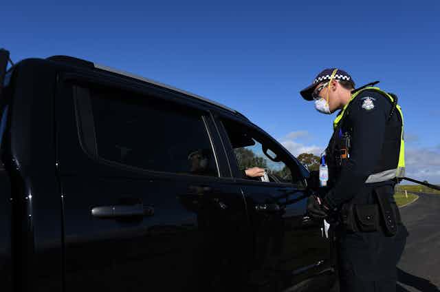 A police officer, wearing a face mask, questions a driver at the border