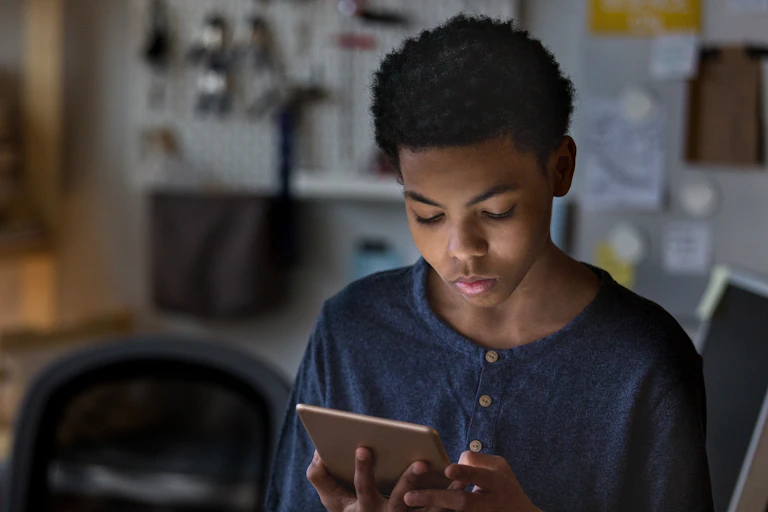 A teen boy plays a game in his room on a digital tablet at night.