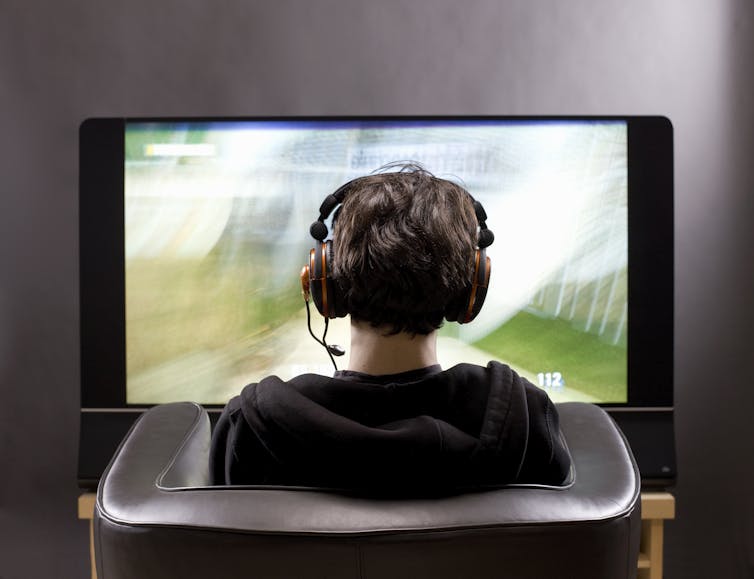 Teenage boy sits in chair playing a video game on a big flat screen.