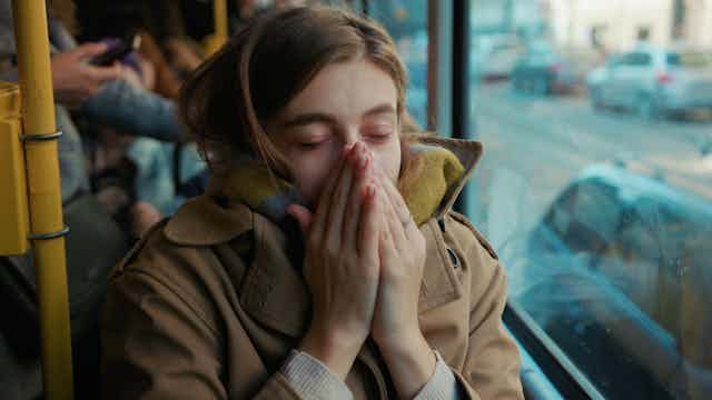 Una chica pálida en un autobús se tapa la cara con las manos para estornudar.