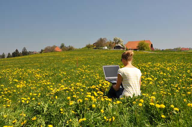 una chica de espaldas trabaja en un ordenador sentada en un campo primaveral con casas al fondo.