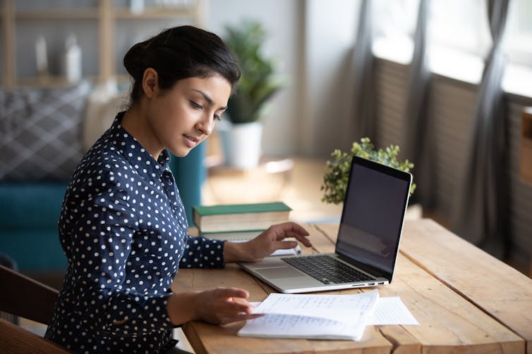 Young woman sitting at desk with laptop and checking notes for assignment