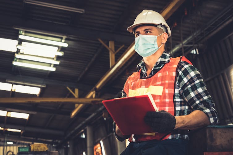 Construction worker wears fluorescent vest, mask and hat.