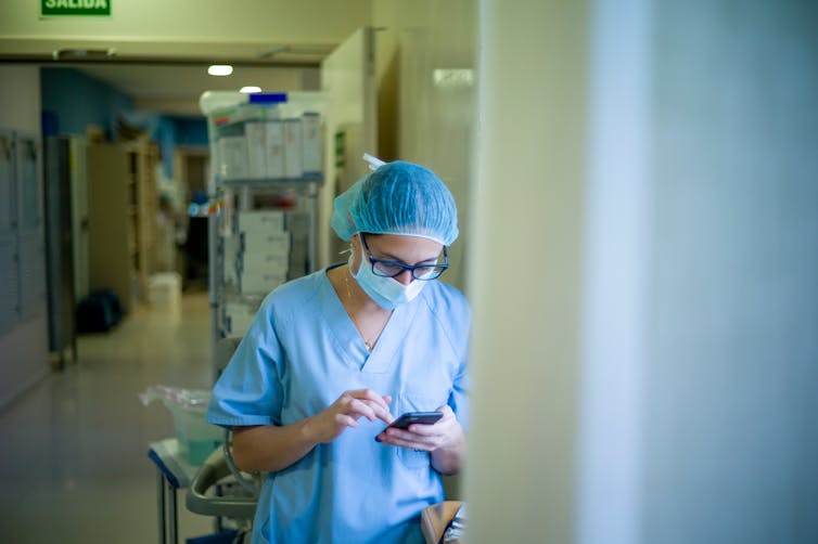 A nurse stands in hospital corridor scrolling on her phone. She is wearing scrubs, a surgical mask and a hairnet.