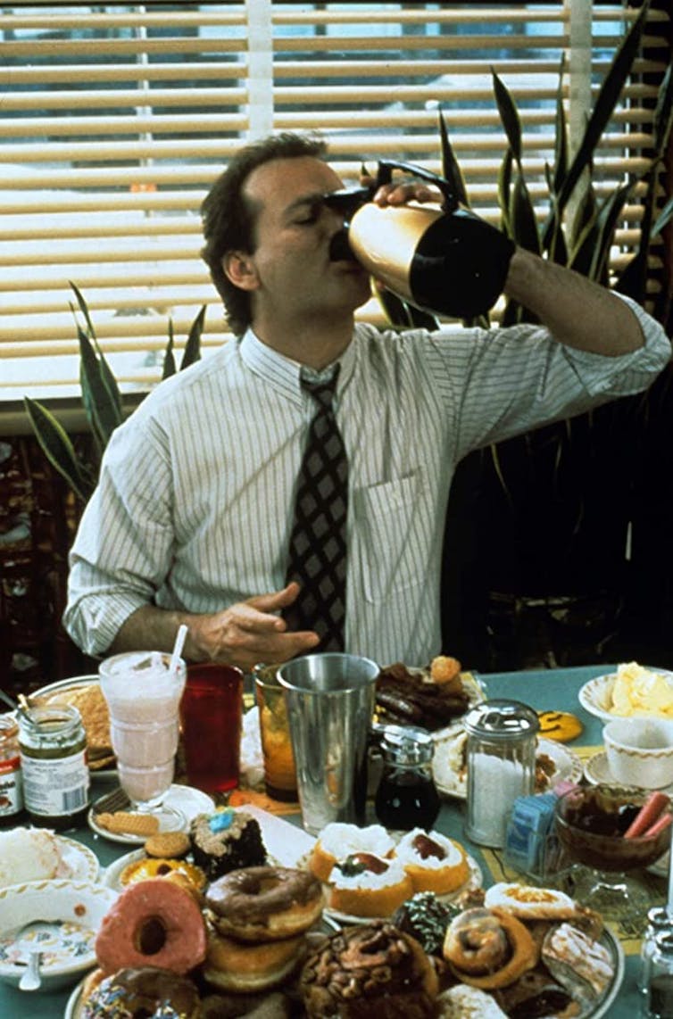 Phil drinks coffee straight from the jug, over a table filled with donuts.