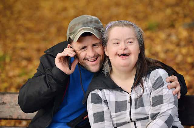 A couple with Down's Syndrome smiles at the camera, golden autumn leaves behind them.