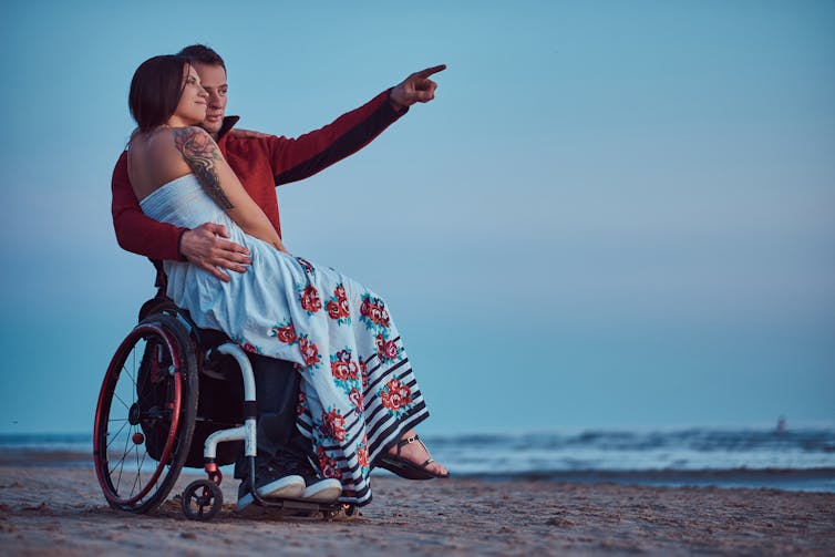 A woman sit on a wheelchair-bound man's lap as he points to something in the distance on a beach.