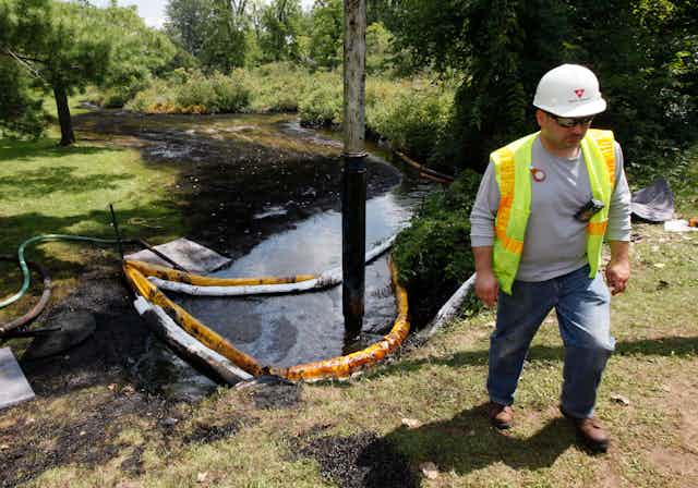 A worker stands on the bank of a creek covered in oil.