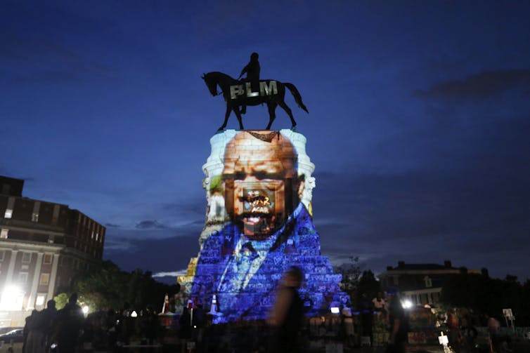 A statue of a man on horseback has a color image of a Black man projected onto its base.