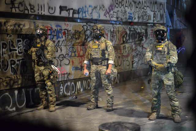 Armed federal officers stand in front of graffiti.