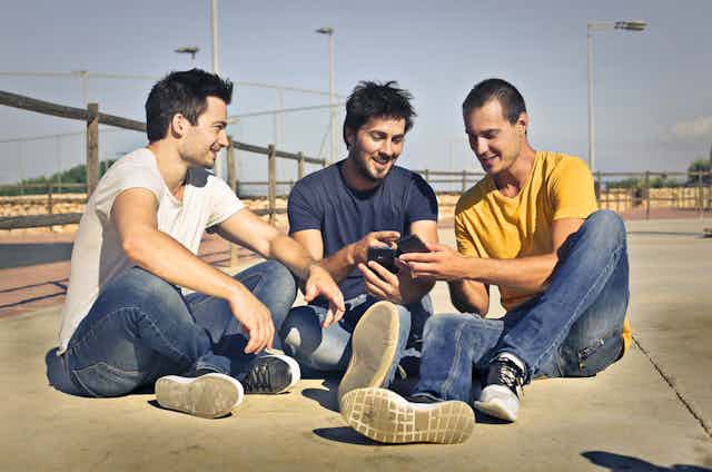 Three young men, smiling and sitting on the ground looking at their mobile phones