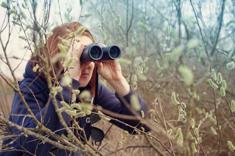 A woman holds binoculars to her eyes among trees