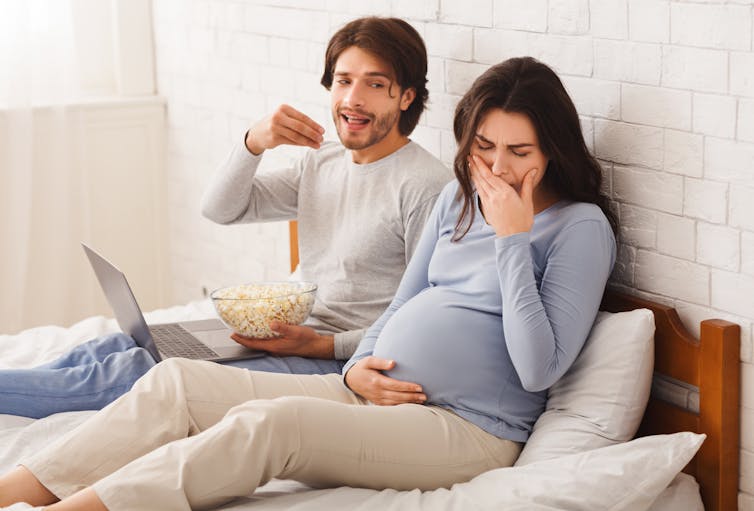 Man eating food on bed while pregnant women covers nose.
