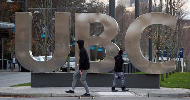 A man on a skateboard and a young woman pass large letters spelling out UBC at the University of British Columbia in Vancouver.