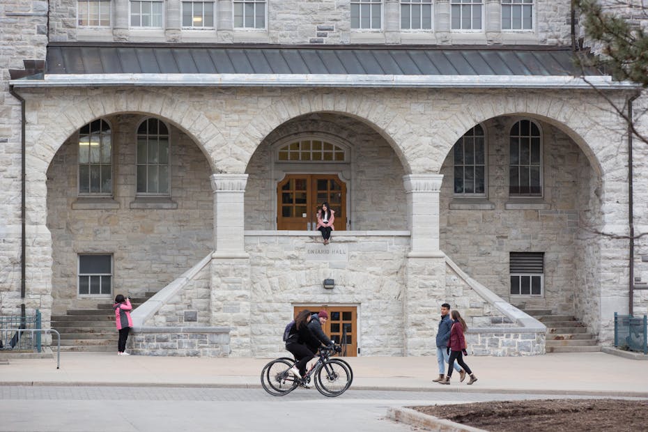 A student on a bicycle and two people pass a stone university building while another student sits on the building's railing.