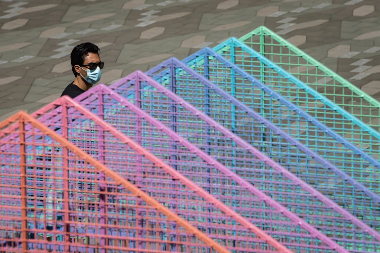 A dark-haired man wearing a blue face mask walks behind a colourful metallic art installation.