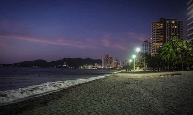 A tropical beach in the evening, with street lamps casting light on the sea.
