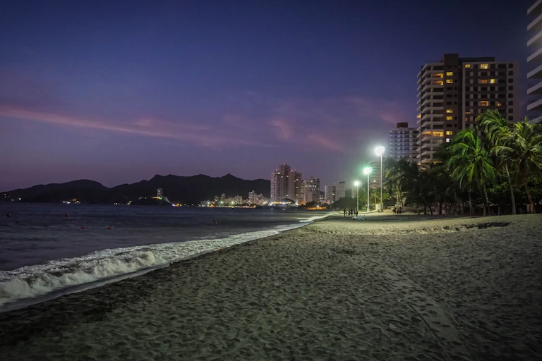 A tropical beach in the evening, with street lamps casting light on the sea.