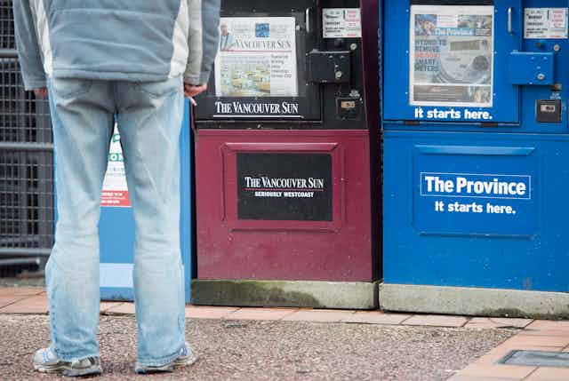 A man stands in front of newspaper boxes.