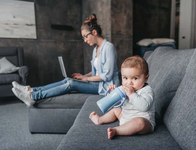 A chubby baby sits on a couch chewing on his sippy cup with his mother behind him on her laptop.