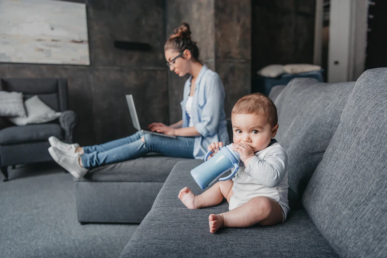 A chubby baby sits on a couch chewing on his sippy cup with his mother behind him on her laptop.