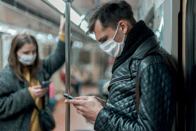 A man in the foreground and a woman in the background stand on a subway train—both are wearing masks and looking at their mobile phones.