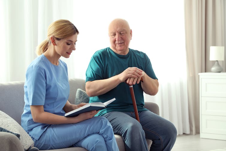 A nurse and a man with a walking stick are seated on a couch. The nurse is reading.