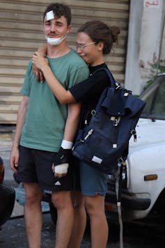 A young man with gauze on his face is hugged by a young woman who is crying.