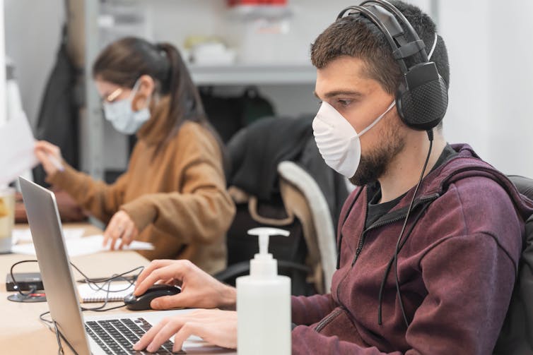 Two office workers wearing surgical masks using laptop.