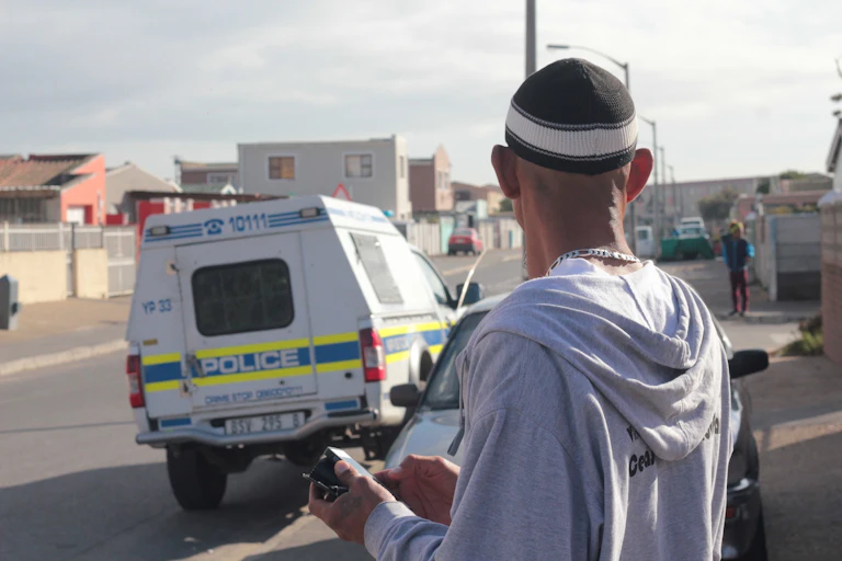 Gang member in skull cap looks at police car parked nearby.