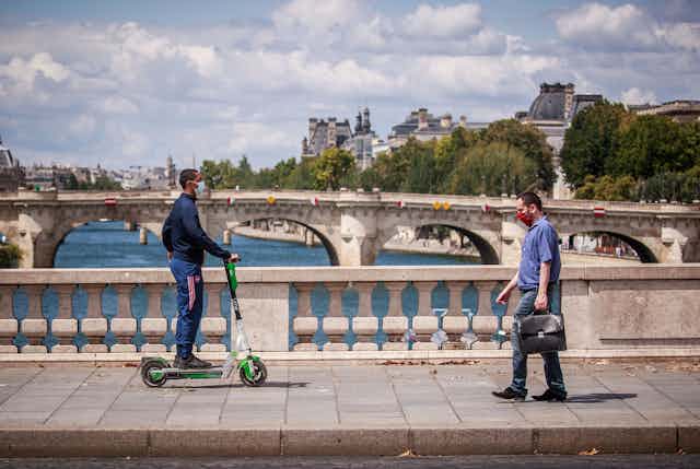 A man with a briefcase and a man on an electric scooter cross a bridge over the Seine, Paris, in opposite directions. 