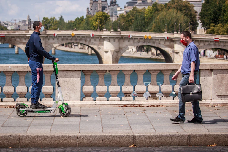 A man with a briefcase and a man on an electric scooter cross a bridge over the Seine, Paris, in opposite directions.