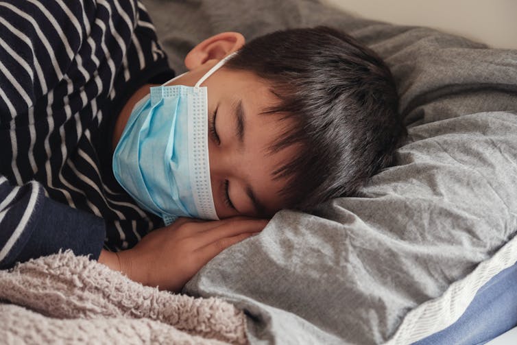 Boy wearing surgical mask is sleeping on grey pillow.