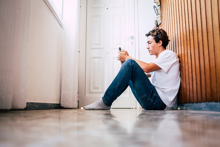 A young man sitting on the floor in a corridor, looking at his phone.