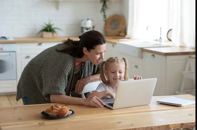 A woman peers over her young daughter's shoulder at a laptop screen set up next to a plate of muffins on a kitchen table.