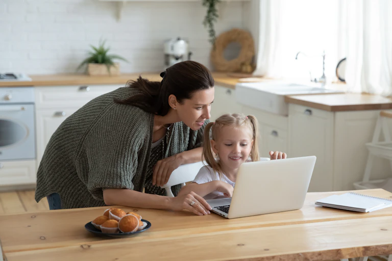 A woman peers over her young daughter's shoulder at a laptop screen set up next to a plate of muffins on a kitchen table.