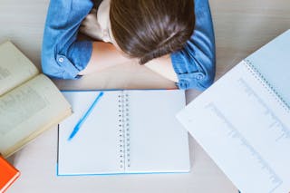 Uma estudante descansa a cabeça, dormindo sobre uma mesa, com livros abertos à sua frente.