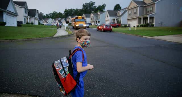 A young boy in a face mask and carrying a backpack looks tentative as he waits by a road in a subdivision as his school bus approaches.