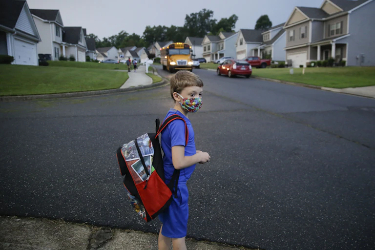 A young boy in a face mask and carrying a backpack looks tentative as he waits by a road in a subdivision as his school bus approaches.