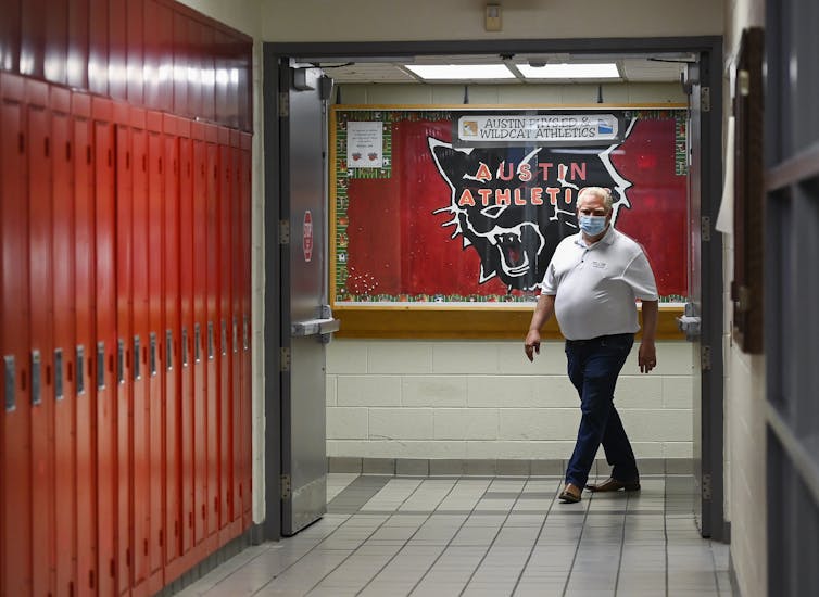 Doug Ford, wearing a mask, walks down a school hallway with a row of red lockers to the left.