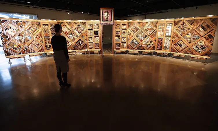 A woman stands in front of the Witness Blanket exhibit at the Canadian Museum for Human Rights. The exhibit features items from the residential schools era including badges and trophies, shoes and a door from one of the schools.