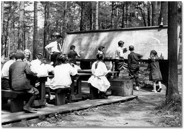 Students sit at desks outdoors in front of a chalkboard.